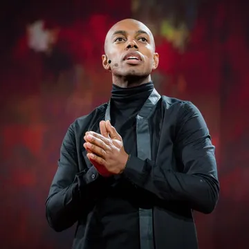 A confident speaker, dressed in black, looking towards the crowd with his hands together, separated visually from a red, textured background via atmospheric perspective.