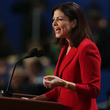 A 3/4 view of a poised speaker, wearing a red suit giving a speech at a lectern. The crowd behind the speaker is blurred and out of focus.